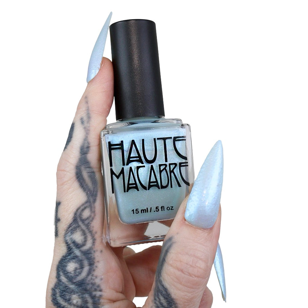 A hand with long, pointed silver-blue manicured nails holds a bottle of Haute Macabre nail polish against a solid white background.
