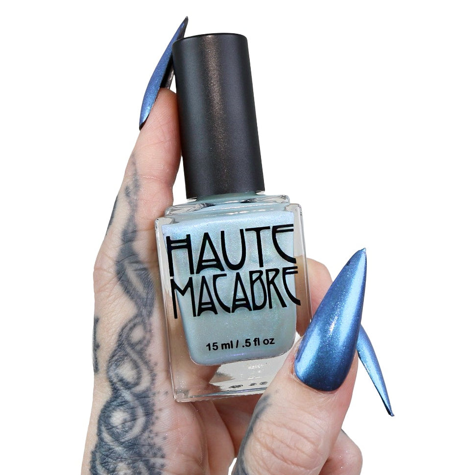 A hand with long, metallic blue manicured nails and a black tattoo holds a bottle of Haute Macabre nail polish against a solid white background.