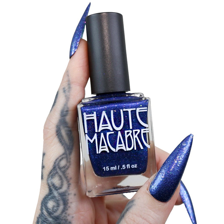 A hand with long, sharp blue manicured nails and tattoos holds a bottle of haute macabre nail polish. The polish is a deep, shimmering royal blue, visible through the clear glass bottle with a black cap.