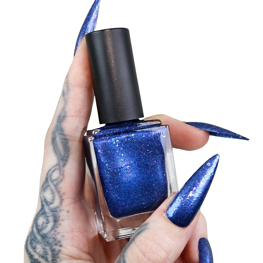 A hand with long, sharp blue stiletto nails and a tattooed forearm holds a bottle of shimmering royal blue nail polish against a plain white background.