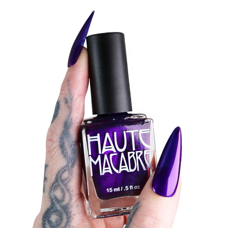 Purple nail polish bottle with 'Haute Macabre' text held by a hand with purple nails on a white background