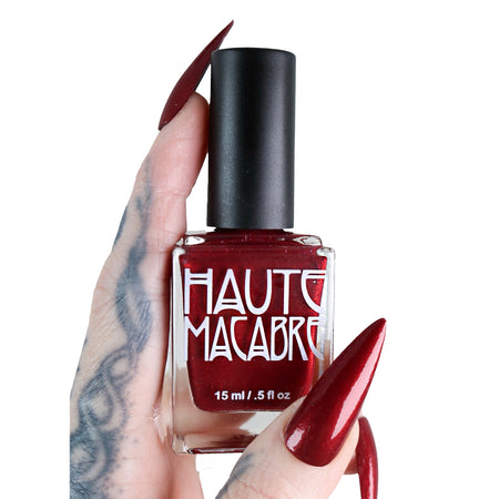 Red nail polish bottle with black cap held by a hand on a white background, featuring 'Haute Macabre' branding.